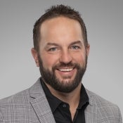 Smiling man wearing a light gray checkered blazer and black shirt against a gray background.