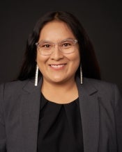 A woman with black hair, earrings, glasses, and a gray blazer smiles in front of a gray background.