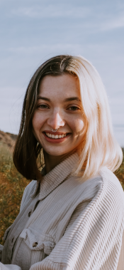 Lynae Carlin, outside and wearing a light colored corduroy shirt, smiles at the camera while highlighted in dramatic sunlight.