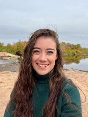 Portrait of a young woman with long curly brown hair wearing a green turtleneck posing in front of a lake