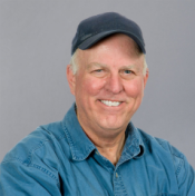 A headshot of a man with white hair, a blue button up shirt, and a baseball cap