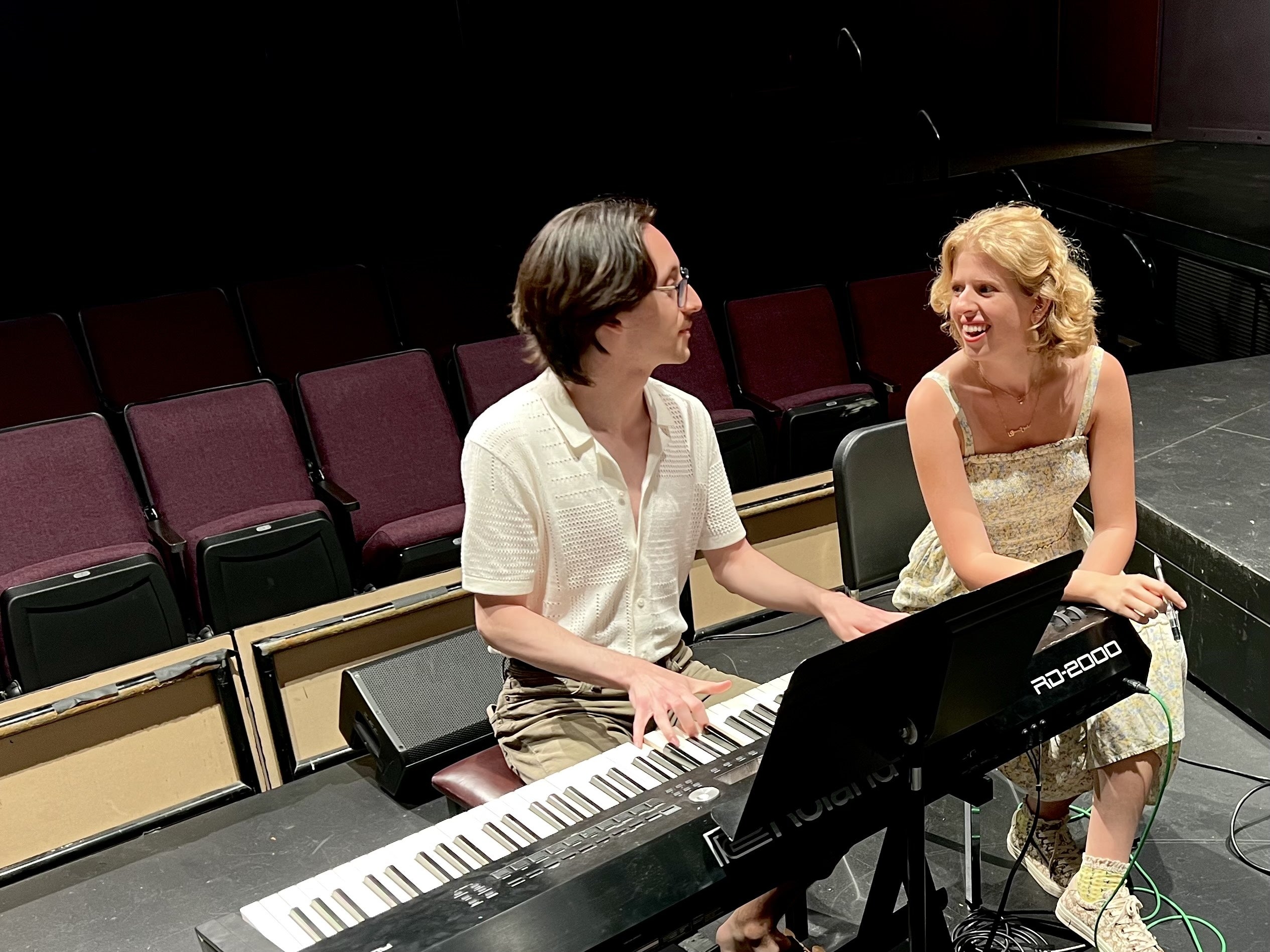 Two people sit behind a piano in a theater looking at each other.