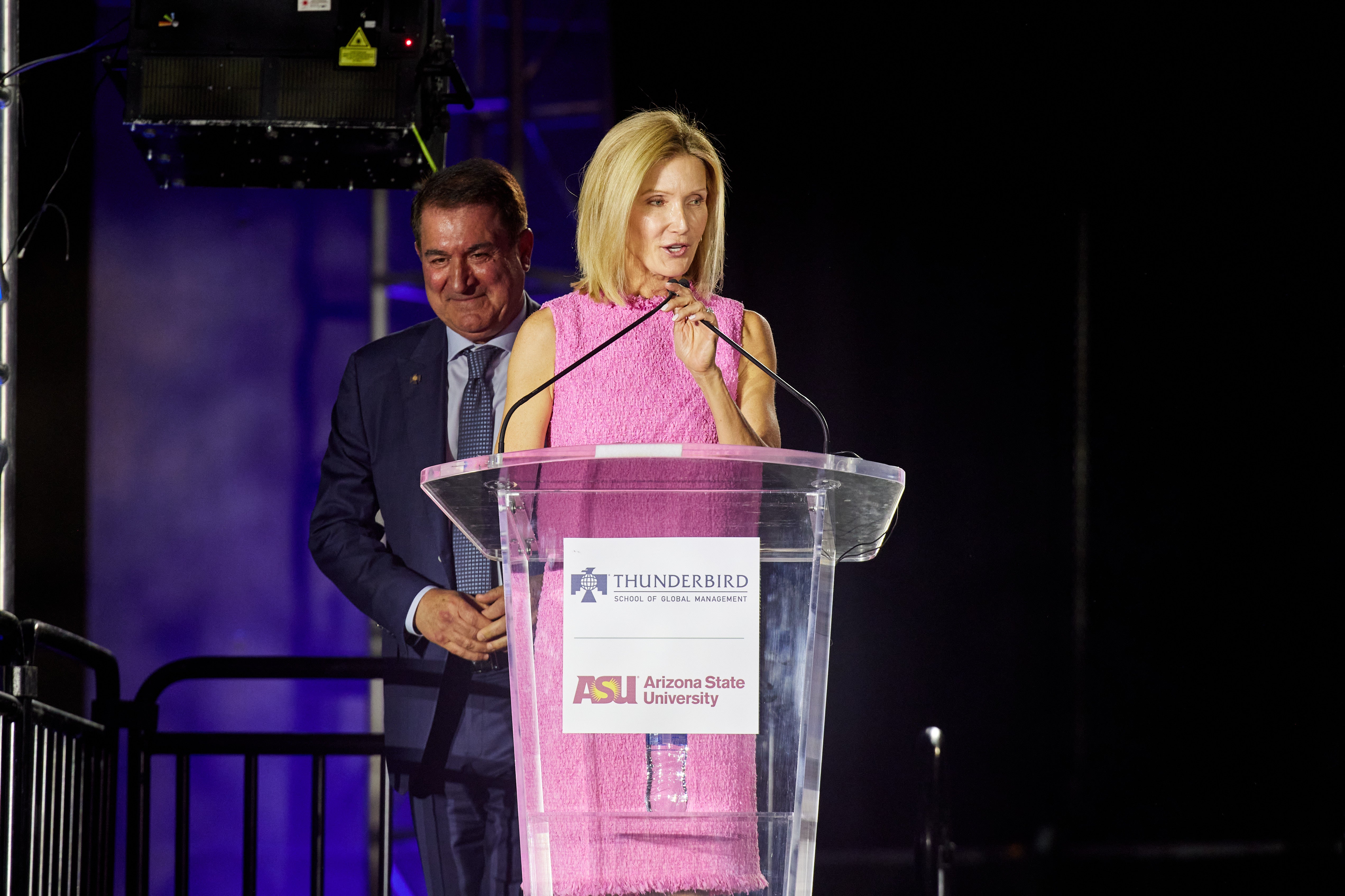 A woman speaks at a lectern with the Thunderbird School logo on it while a man stands behind her