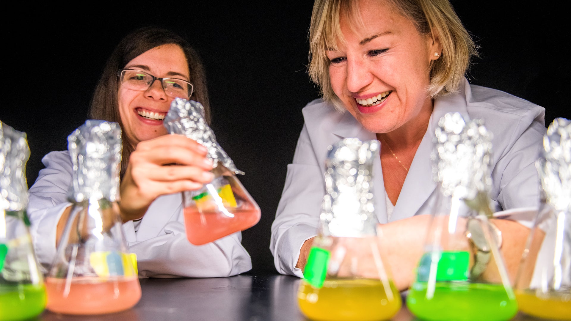 Two women in white coats sit at a table with beakers. 