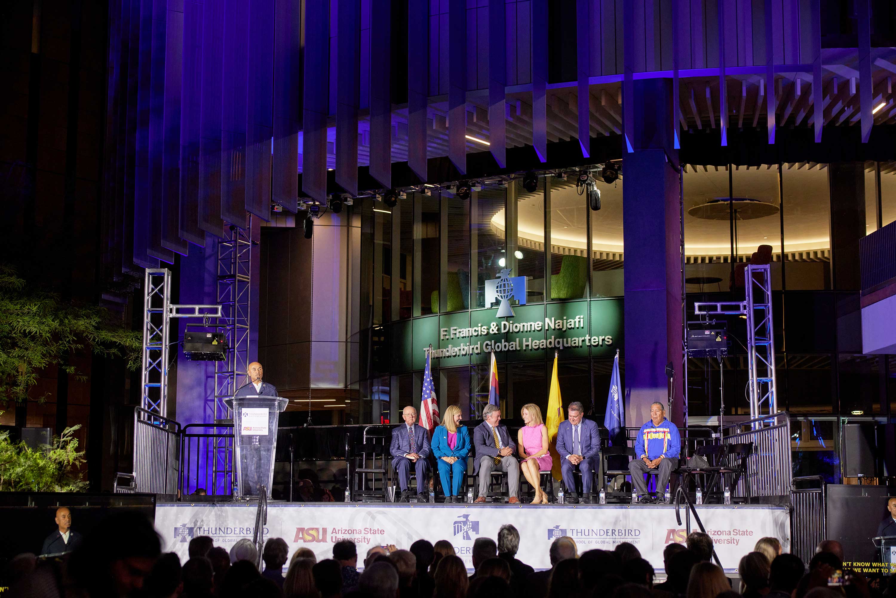 Man speaking at lectern with people sitting nearby on stage during building naming event